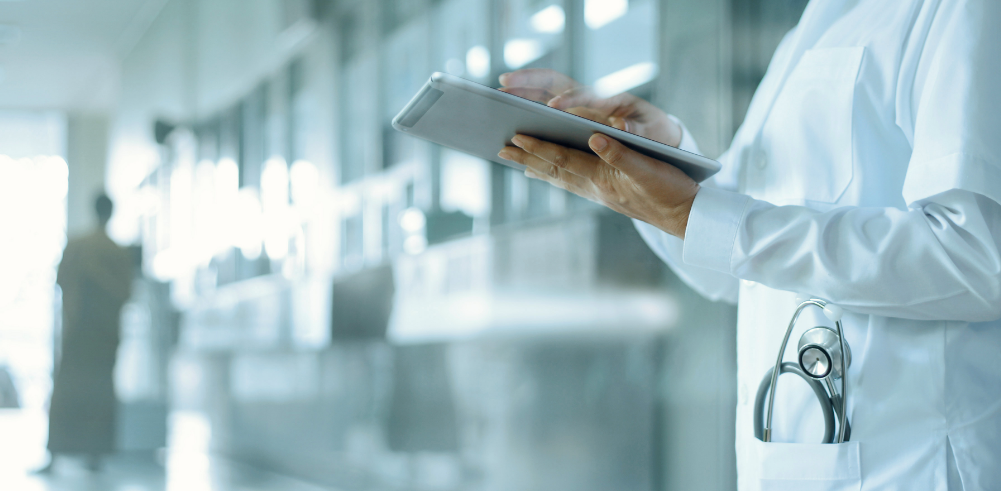 Doctor in a white coat with a stethoscope using a tablet in a bright hospital corridor