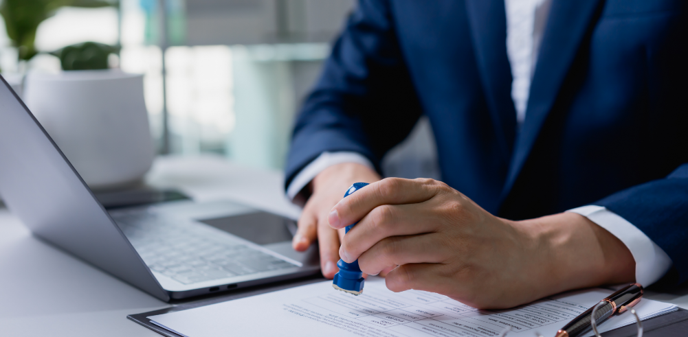 Person in a suit stamping a document at a desk beside a laptop and glasses