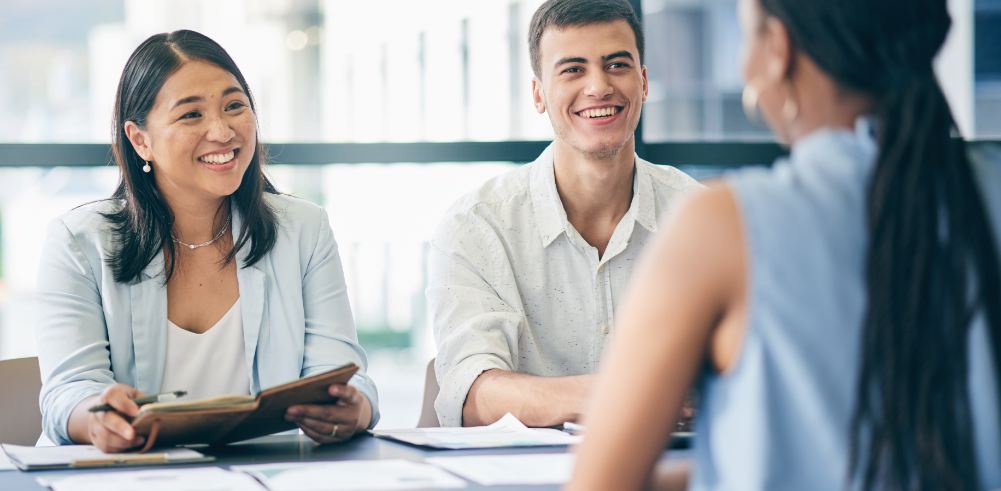 Two smiling colleagues in a meeting with a third person seen from behind, documents on the table
